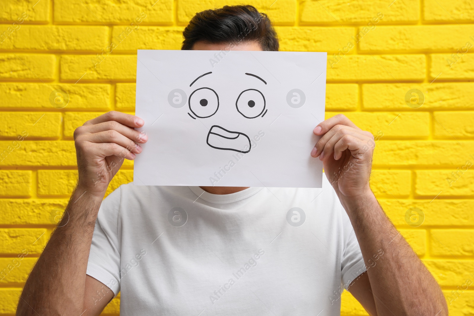 Photo of Man hiding emotions using card with drawn scared face near yellow brick wall