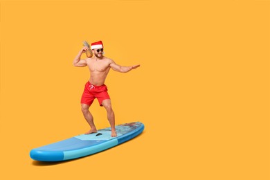 Happy man in Santa hat with pineapple posing on SUP board against orange background. Space for text