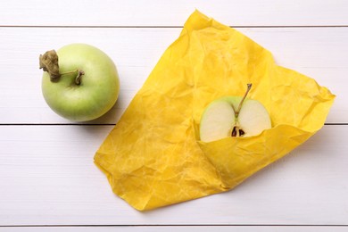 Photo of Half of apple in yellow beeswax food wrap on white wooden table, top view