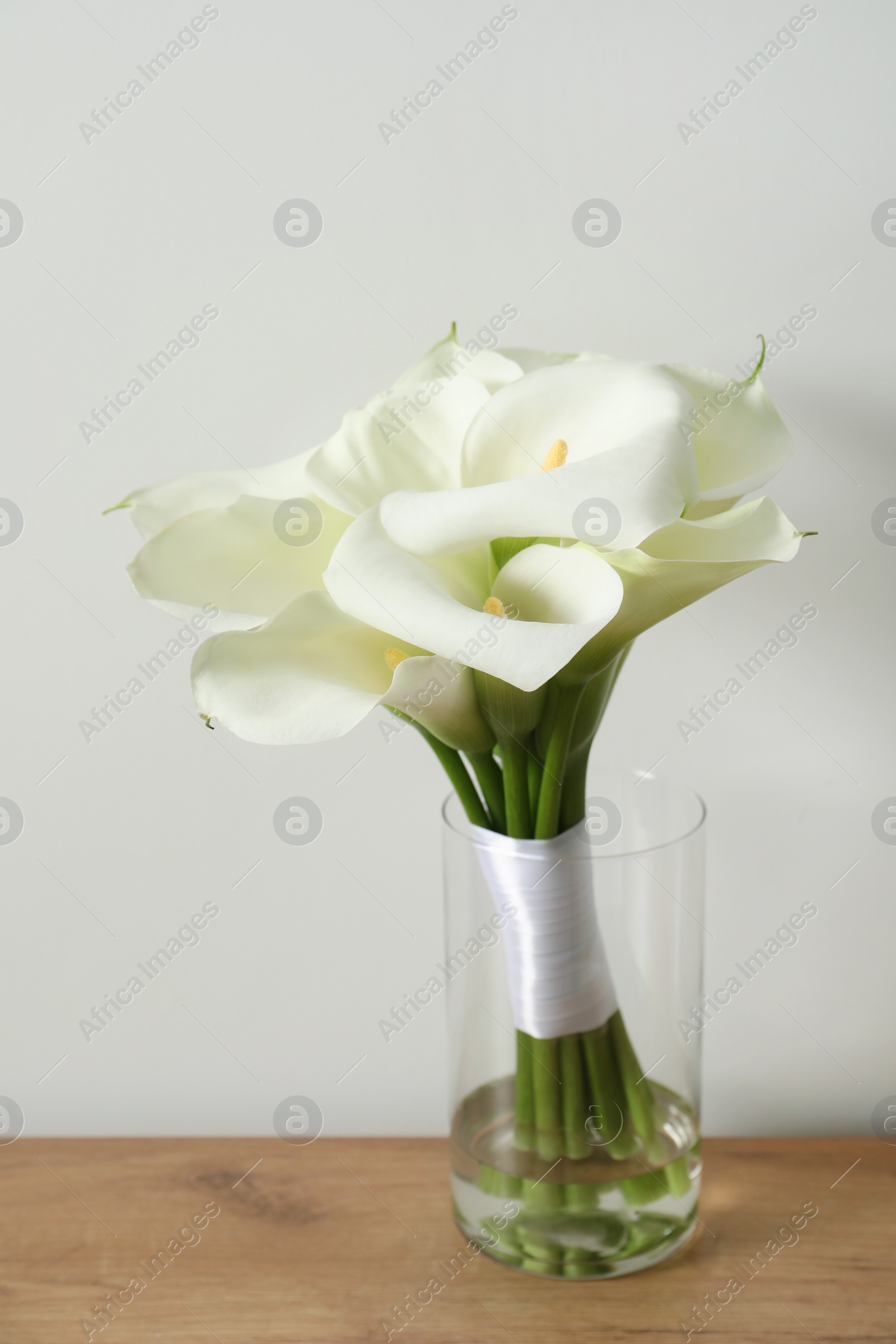 Photo of Beautiful calla lily flowers tied with ribbon on wooden table near white wall