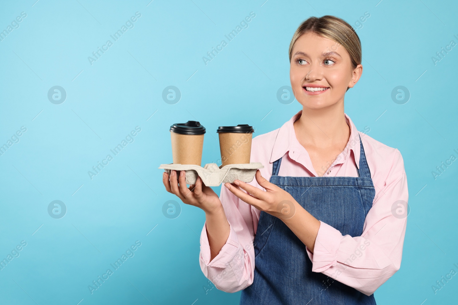 Photo of Beautiful young woman in denim apron with cups of coffee on light blue background. Space for text