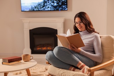 Young woman reading book in armchair near fireplace at home