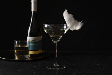Photo of Cocktail with tasty cotton candy and bottle of alcohol drink on dark textured table against black background