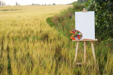 Wooden easel with blank canvas and painting equipment in field. Space for text