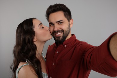 Photo of Woman kissing her smiling boyfriend while he taking selfie on grey background