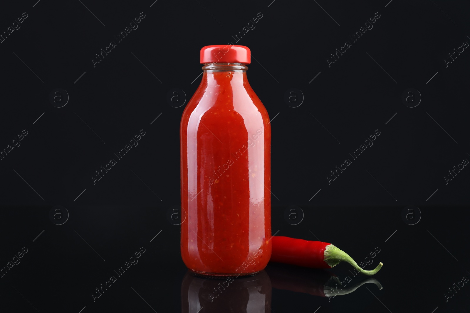 Photo of Spicy chili sauce in bottle and pepper against dark background