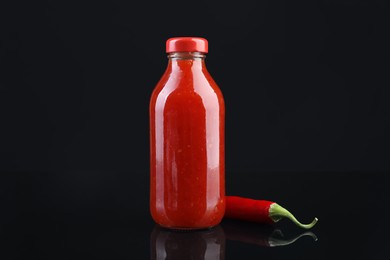 Photo of Spicy chili sauce in bottle and pepper against dark background