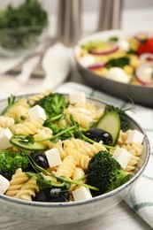 Photo of Bowl of delicious pasta with cucumber, olives, broccoli and cheese on white wooden table, closeup