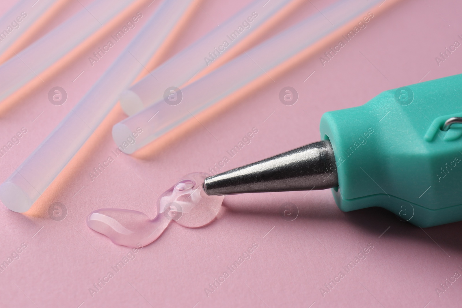 Photo of Melted glue dripping out of hot gun nozzle near sticks on pink background, closeup