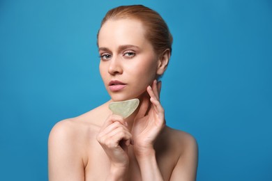 Photo of Beautiful young woman doing facial massage with gua sha tool on blue background