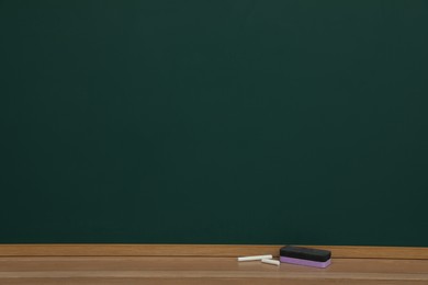 Pieces of chalk and duster on wooden table near greenboard space for text