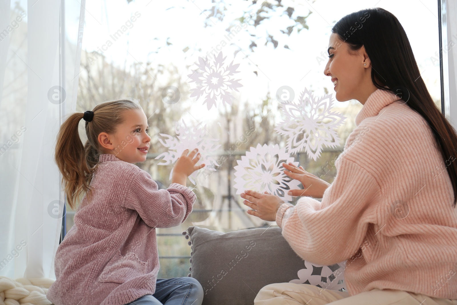 Photo of Mother and daughter decorating window with paper snowflakes at home