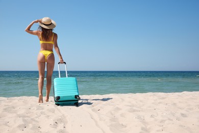 Photo of Woman in bikini with suitcase on sandy beach near sea, back view