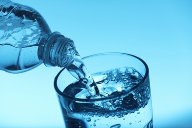 Pouring water from bottle into glass on light blue background, closeup