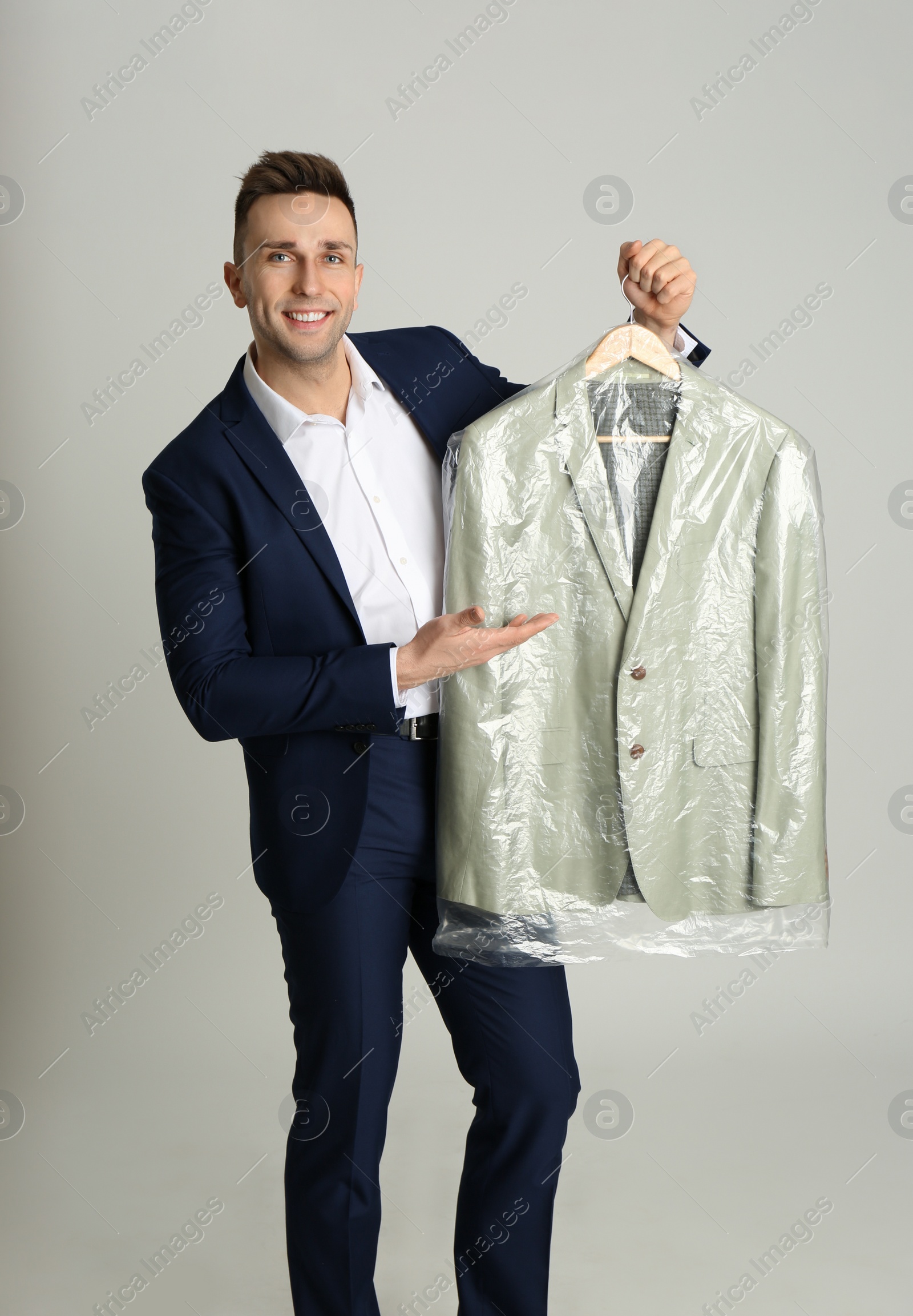 Photo of Man holding hanger with jacket in plastic bag on light grey background. Dry-cleaning service