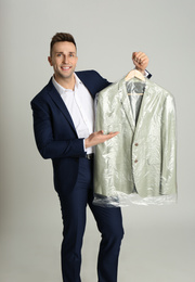 Man holding hanger with jacket in plastic bag on light grey background. Dry-cleaning service