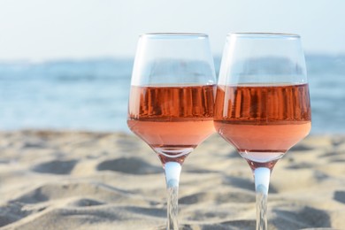 Photo of Glasses of tasty rose wine on sand near sea, closeup. Space for text
