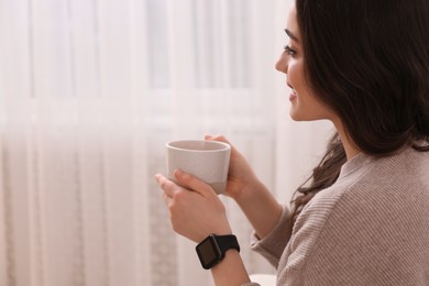 Young woman with smart watch and cup of drink at home, space for text