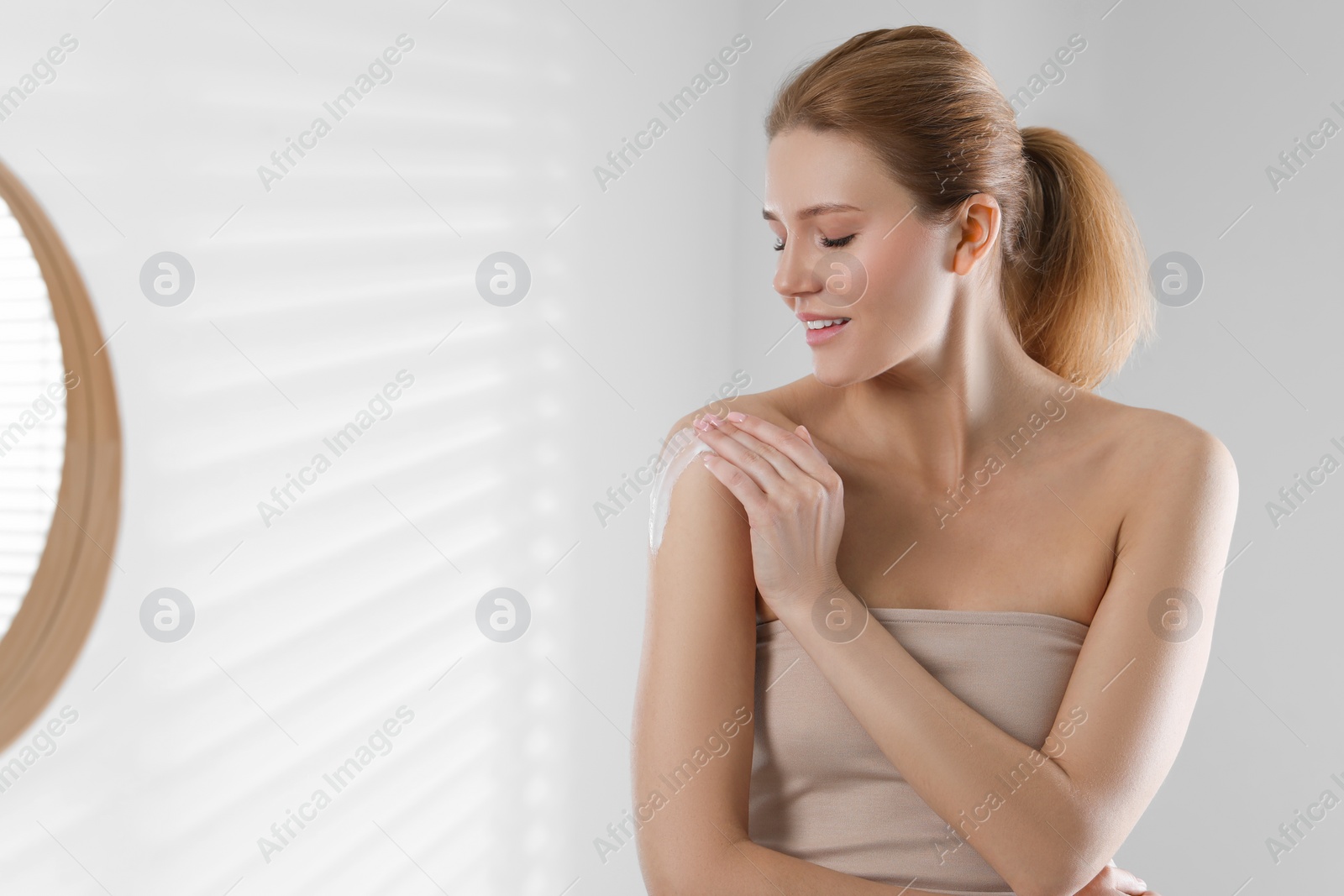 Photo of Beautiful woman applying body cream onto shoulder indoors, space for text