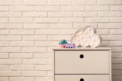 Photo of Chest of drawers with lamp and toys near white brick wall. Space for text