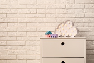 Chest of drawers with lamp and toys near white brick wall. Space for text