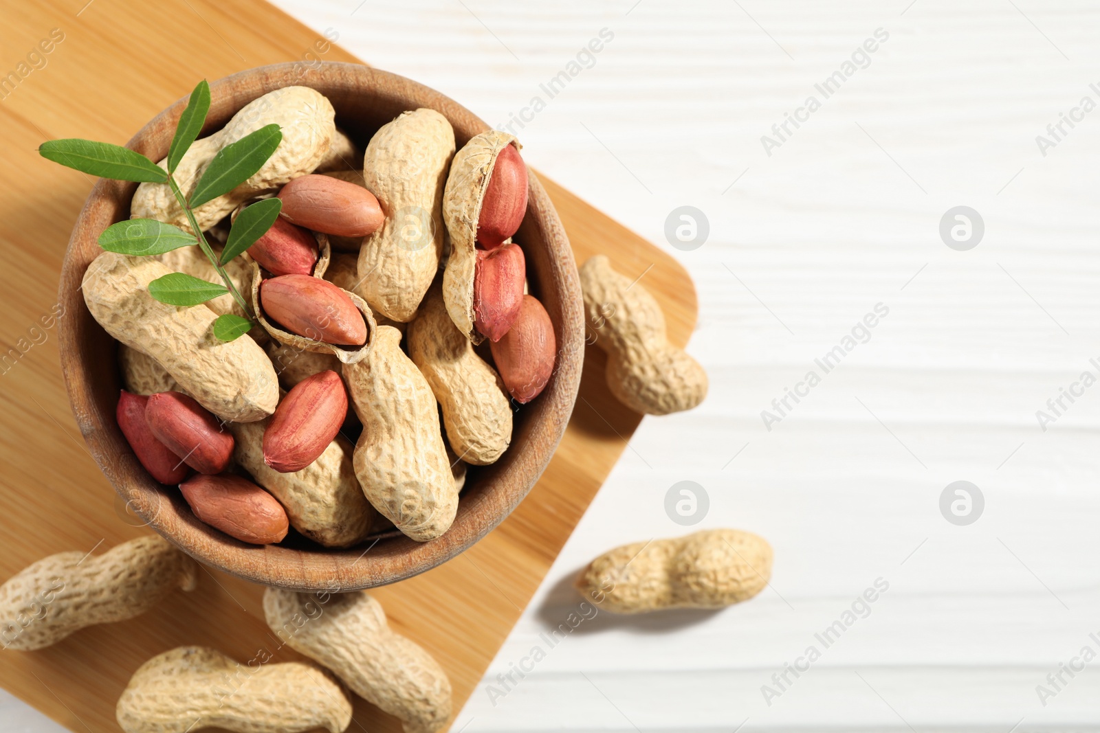 Photo of Fresh unpeeled peanuts in bowl and twig on white wooden table, top view. Space for text