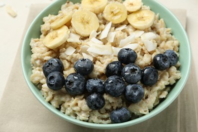 Photo of Tasty oatmeal with banana, blueberries, coconut flakes and honey served in bowl on beige table, closeup