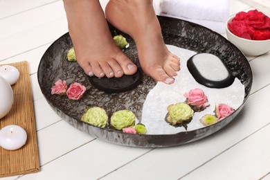 Photo of Woman soaking her feet in plate with water, stones and flowers on white wooden floor, closeup. Pedicure procedure