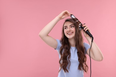 Beautiful young woman using curling hair iron on pink background, space for text