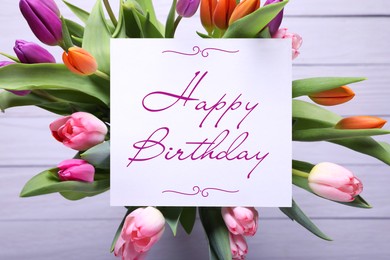 Beautiful bouquet of tulip flowers with Happy Birthday card on white wooden background, top view