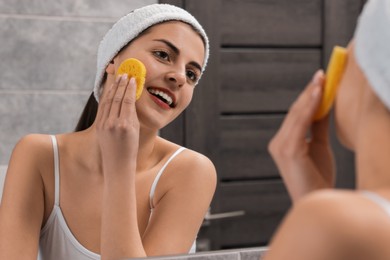 Photo of Young woman with headband washing her face using sponge near mirror in bathroom