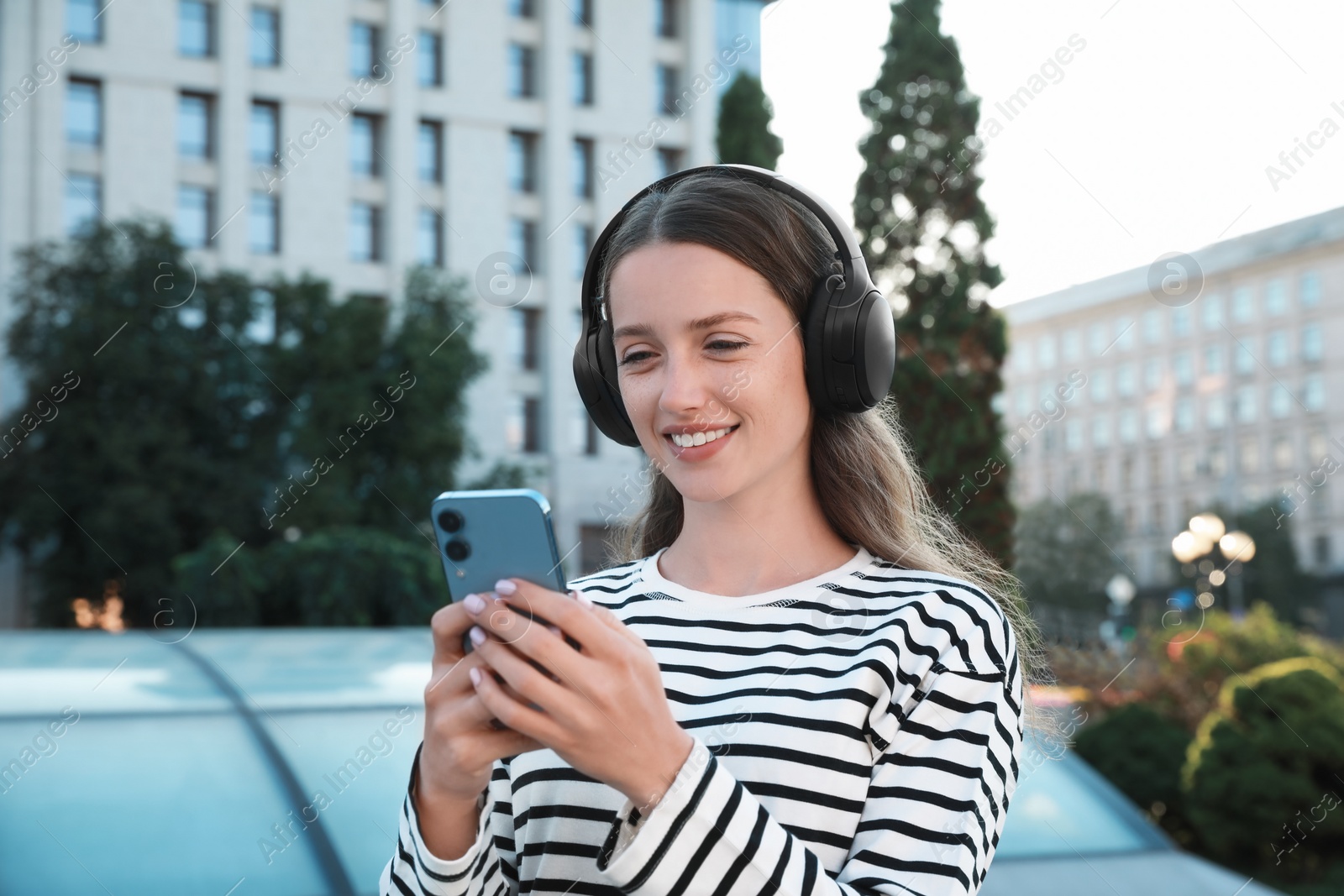 Photo of Smiling woman in headphones using smartphone on city street