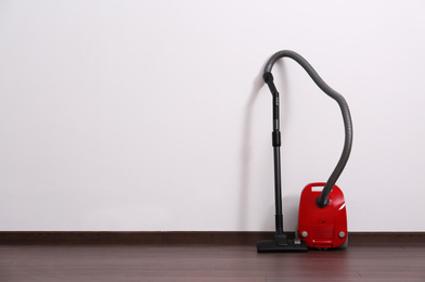 Modern red vacuum cleaner on wooden floor near white wall, space for text