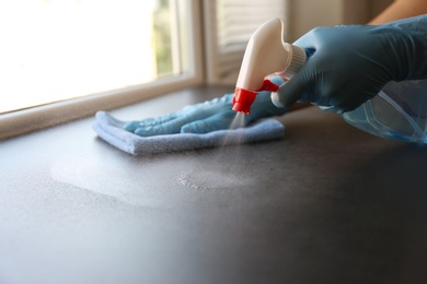 Photo of Woman in gloves cleaning grey window sill with rag and detergent indoors, closeup