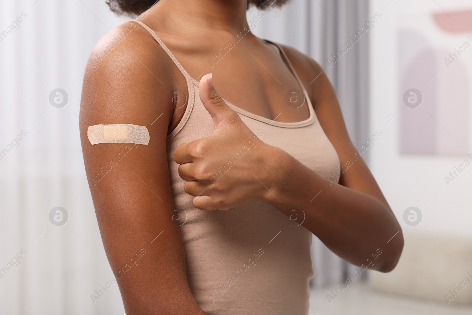 Photo of Young woman with adhesive bandage on her arm after vaccination showing thumb up indoors, closeup