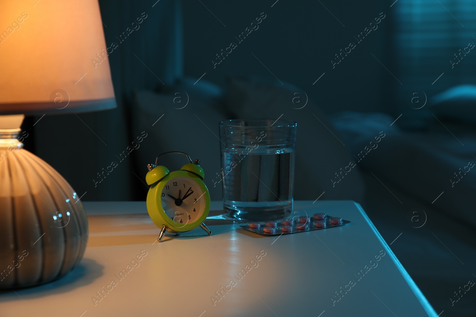 Photo of Insomnia treatment. Glass of water, pills and alarm clock on bedside table in bedroom at night