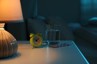 Insomnia treatment. Glass of water, pills and alarm clock on bedside table in bedroom at night