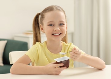 Cute little girl with tasty yogurt at white table indoors