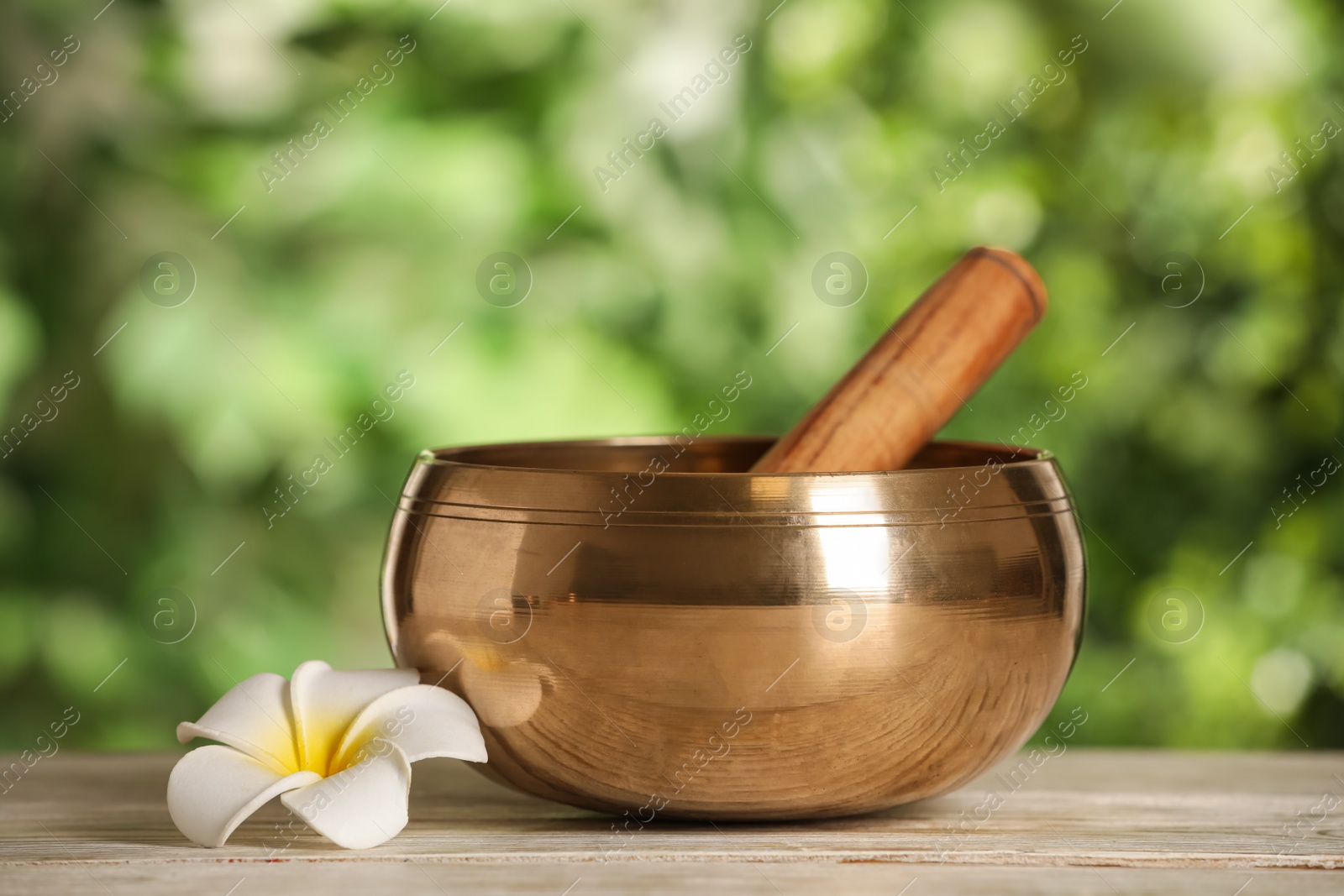 Photo of Golden singing bowl, mallet and flower on white wooden table outdoors