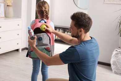 Photo of Happy father putting apple into daughter`s backpack at home. Preparing to school