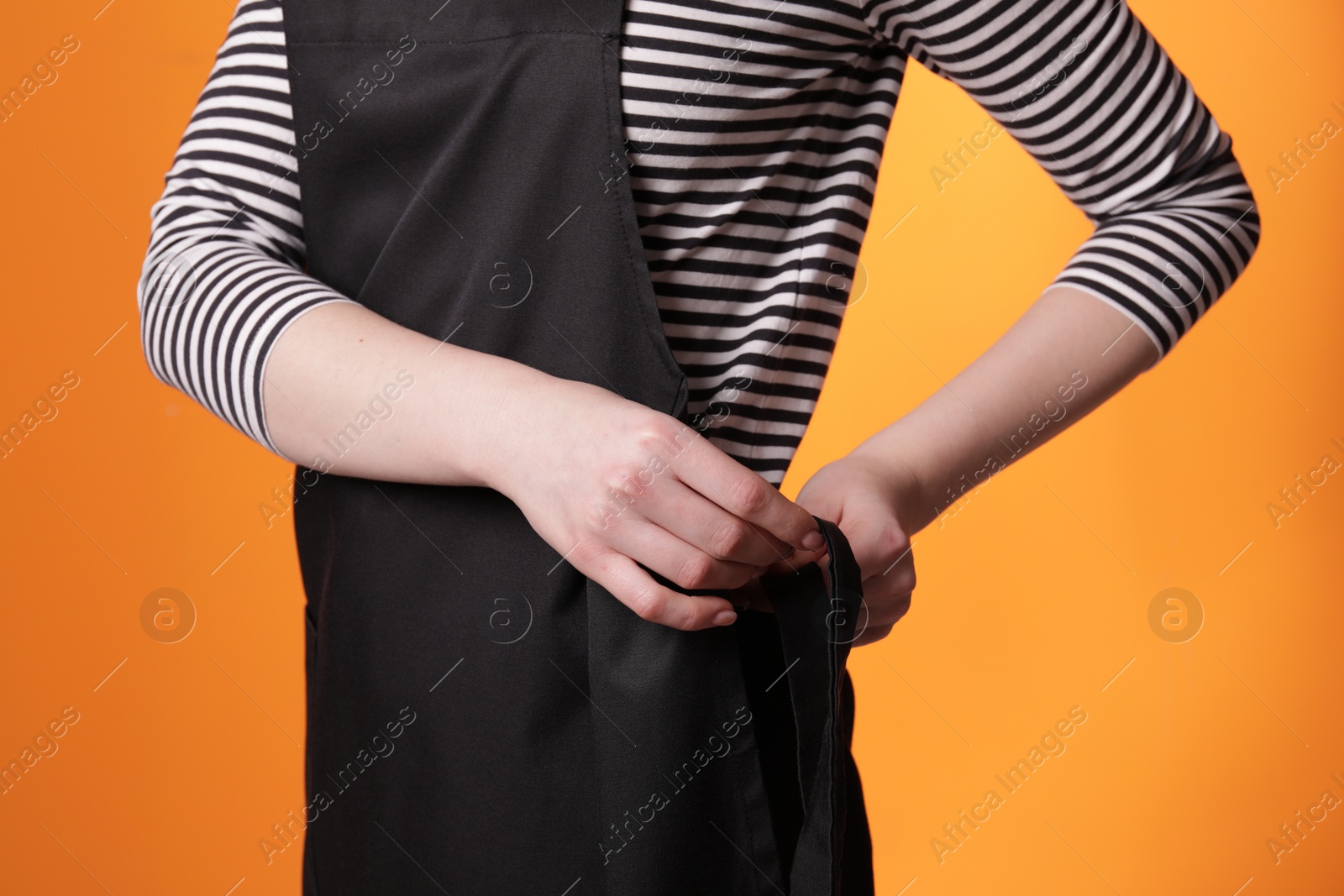 Photo of Woman putting on black apron against orange background, closeup