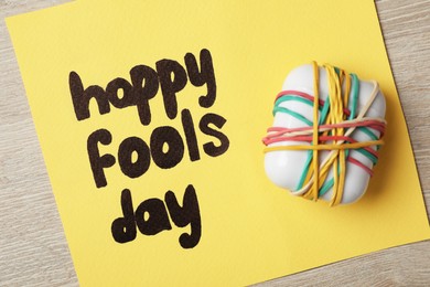 Photo of Earphones case covered with rubber bands and Happy Fools' Day note on wooden background, top view