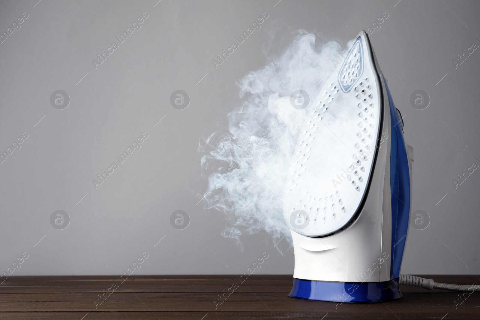 Image of Modern iron with steam on wooden table against light grey background, space for text