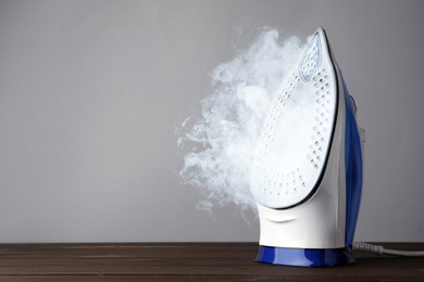 Modern iron with steam on wooden table against light grey background, space for text