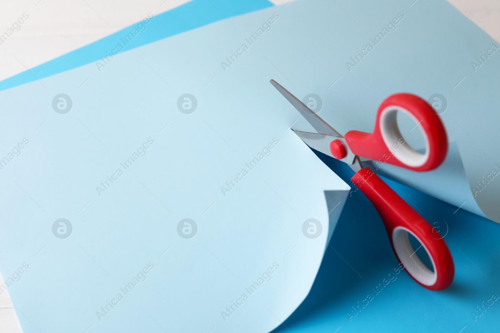 Photo of Red scissors cutting light blue paper on white background, closeup