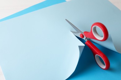 Photo of Red scissors cutting light blue paper on white background, closeup