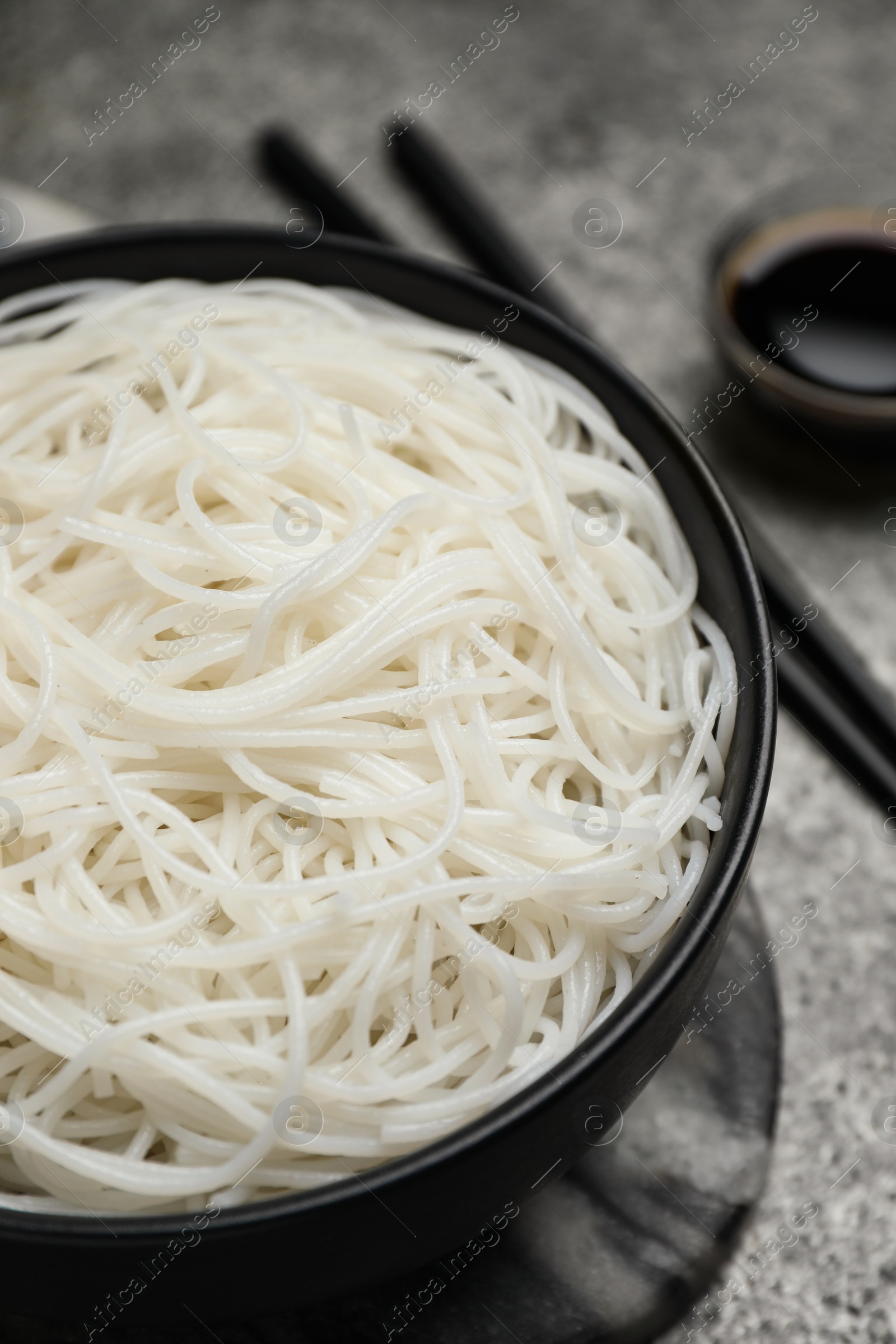 Photo of Bowl with cooked rice noodles, soy sauce and chopsticks on grey table, closeup