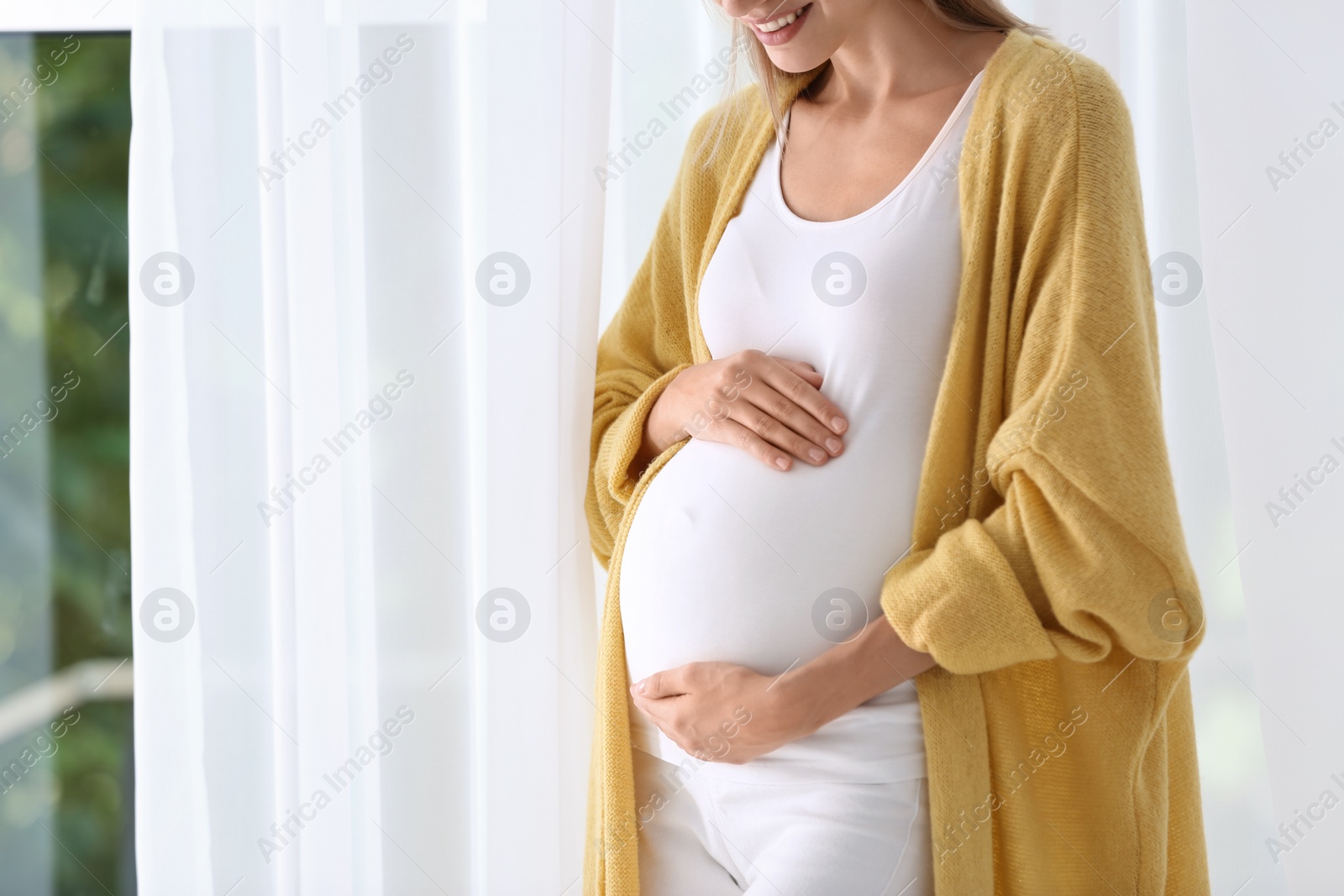 Photo of Pregnant woman standing near window at home, closeup