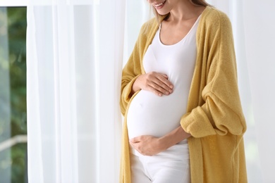 Pregnant woman standing near window at home, closeup