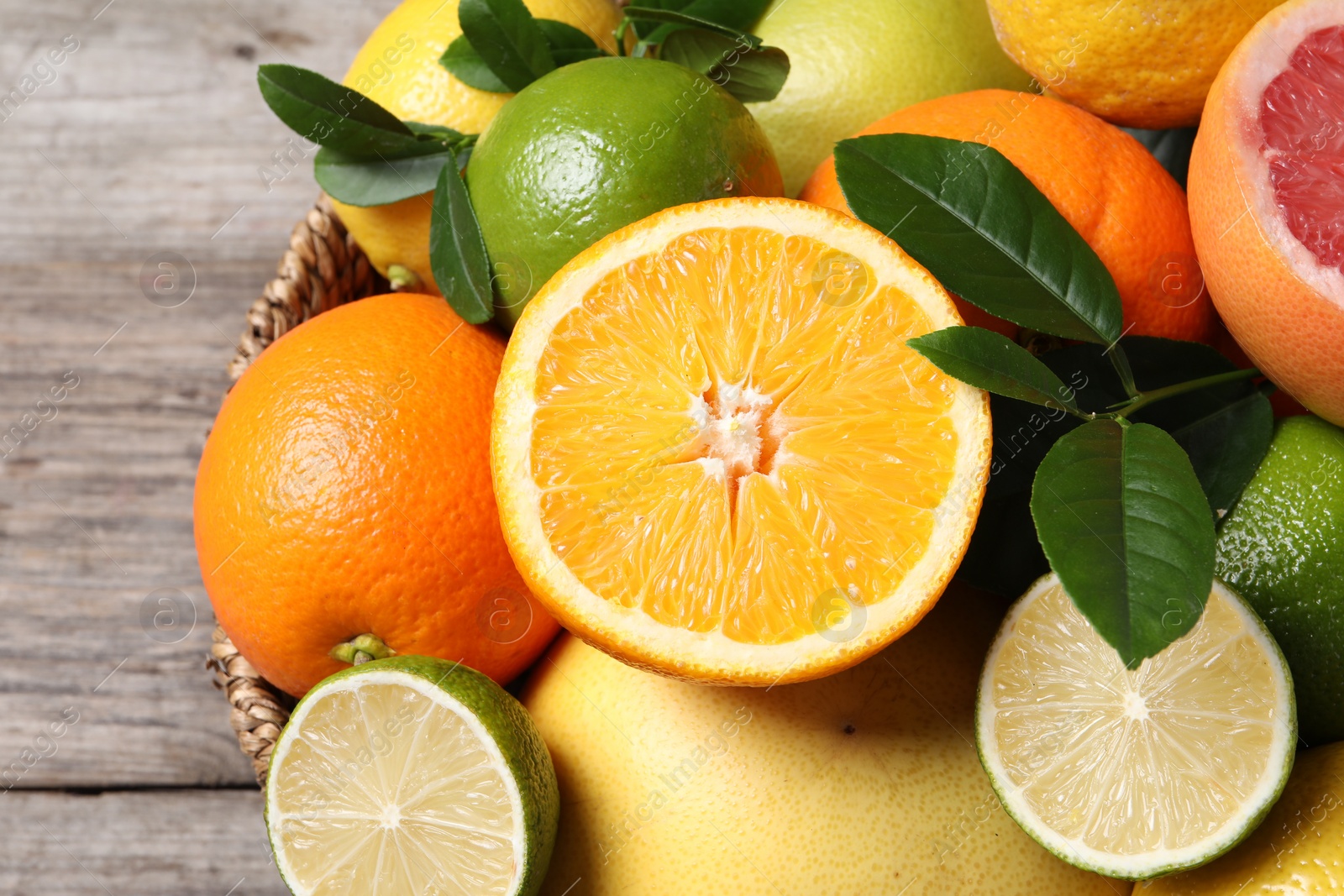 Photo of Different fresh citrus fruits and leaves in wicker basket on wooden table, closeup